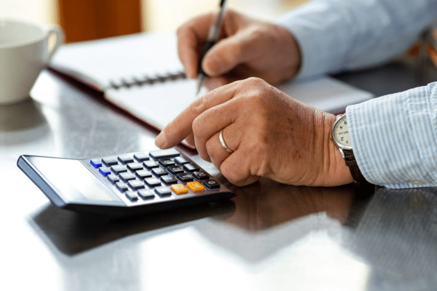 An unrecognizable businessman using a calculator and writing in a notepad at home on a counter to ensure he's making timely payments.